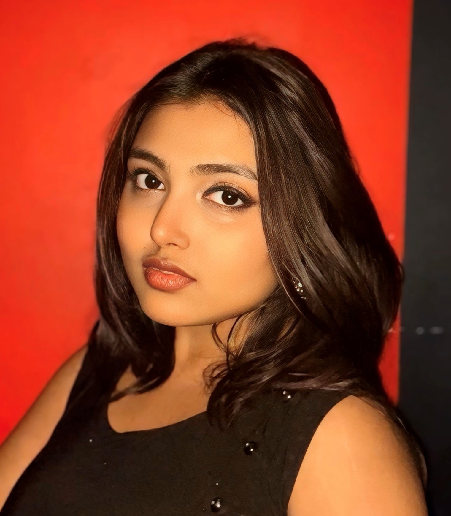 Ankita is standing in front of a red wall. She has long brown hair, she is wearing a black vest, standing at 45 degrees to the camera looking straight ahead