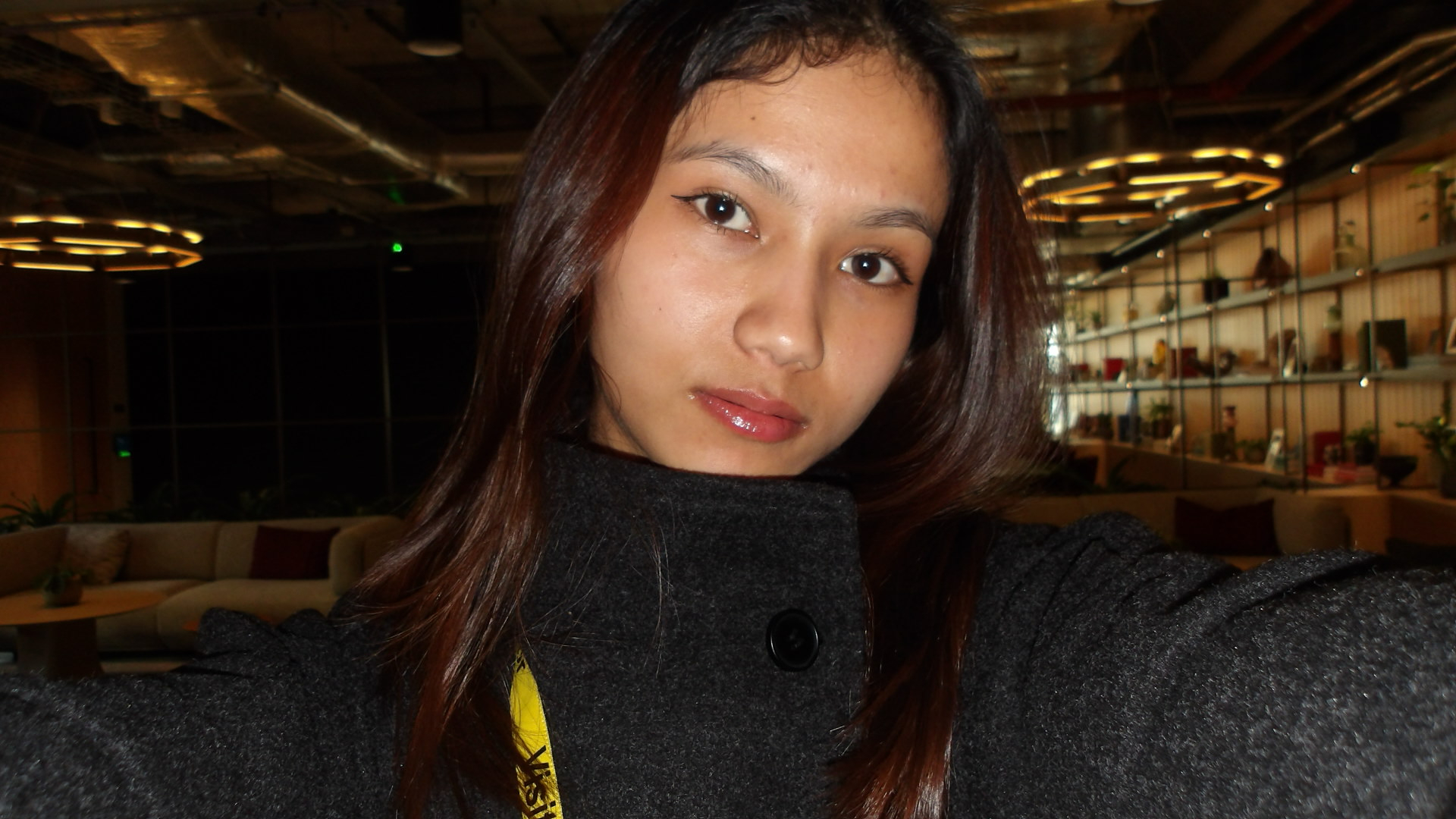 Kritika standing in a dim room with sofas and shelves in the background. She has brown hair and brown eyes. Kritika is wearing a black jacket and looking directly at a camara