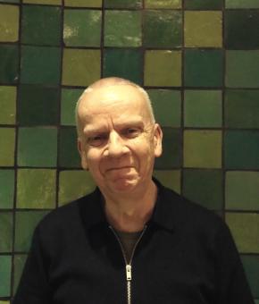 Simeon has short grey hair and is standing in front of a wall covered in green tiles. 