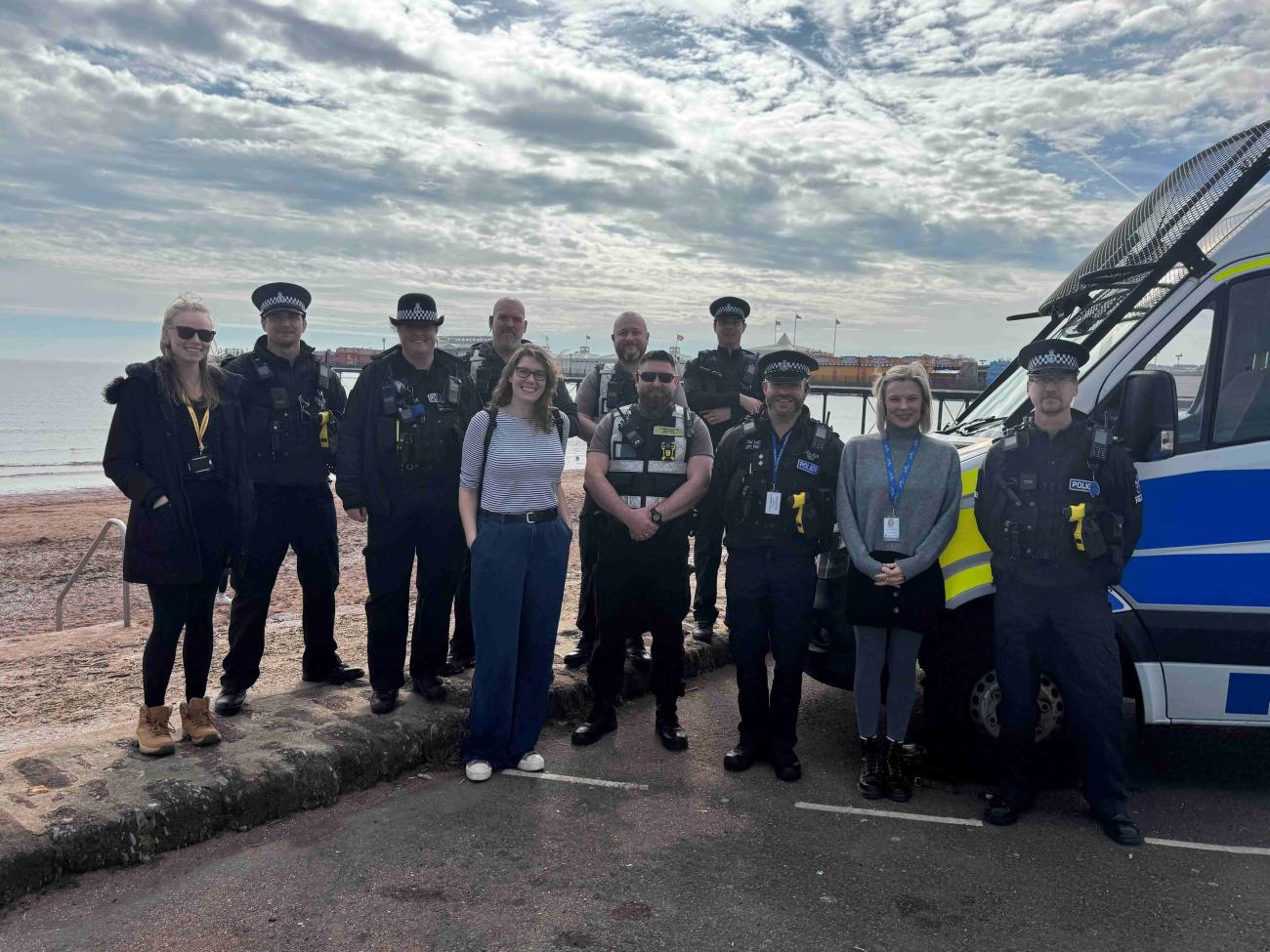Image shows a group of police and NW staff doing a leaflet drop in Paignton, they are in a group of about 11 people posing by a police van next to the sea