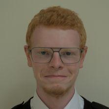 Roke has short red hair and a beard and he is wearing glasses. 