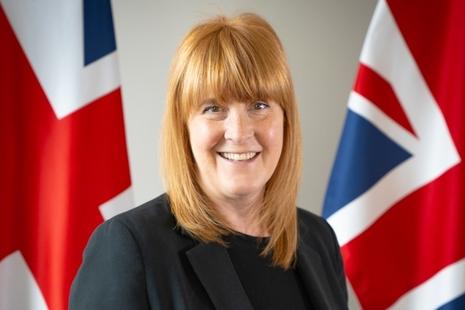 Sarah has red hair, and she is smiling at the camera. She wears a dark suit and is standing in front of two Union Jack flags