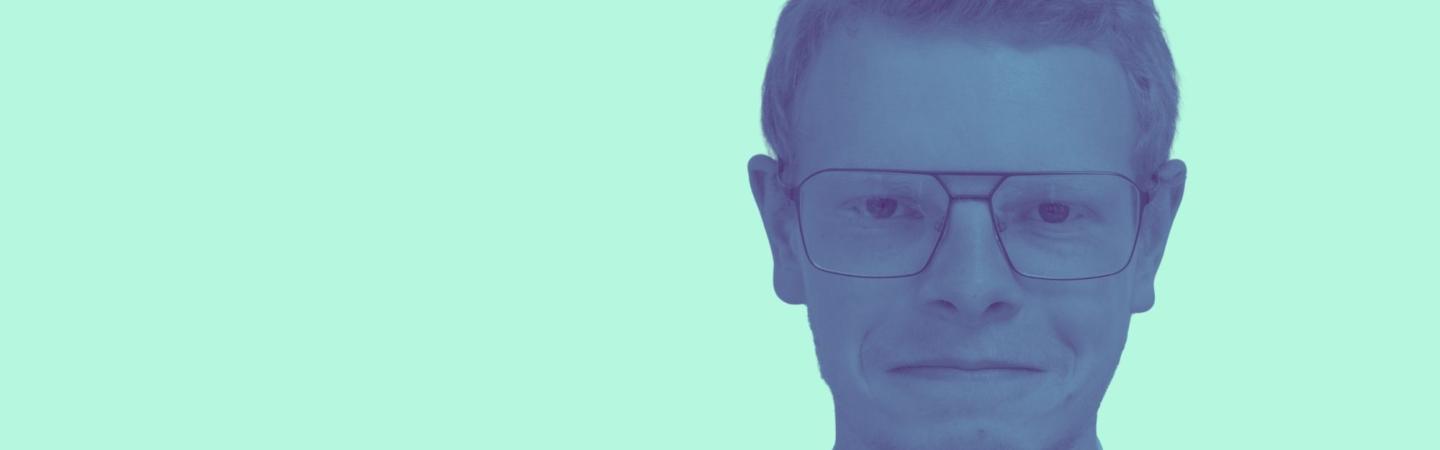 Image shows Roke smiling directly at the camera, he has short red hair and glasses. 